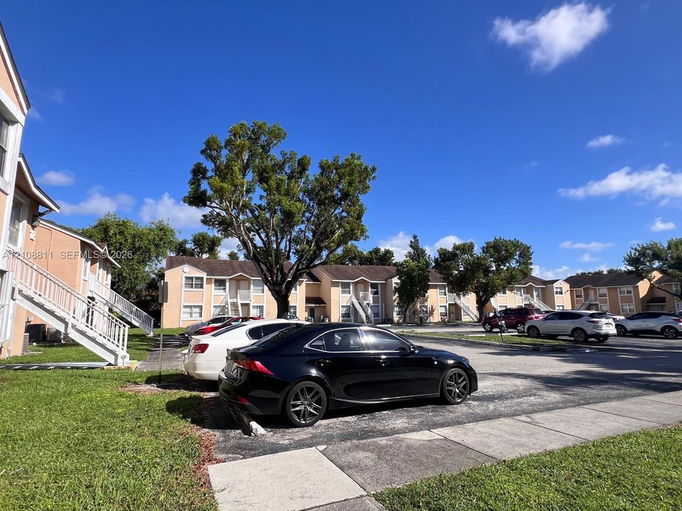 Multi-story residential complex featuring stucco siding, gabled rooflines, exterior staircases, asphalt parking area, and mature trees