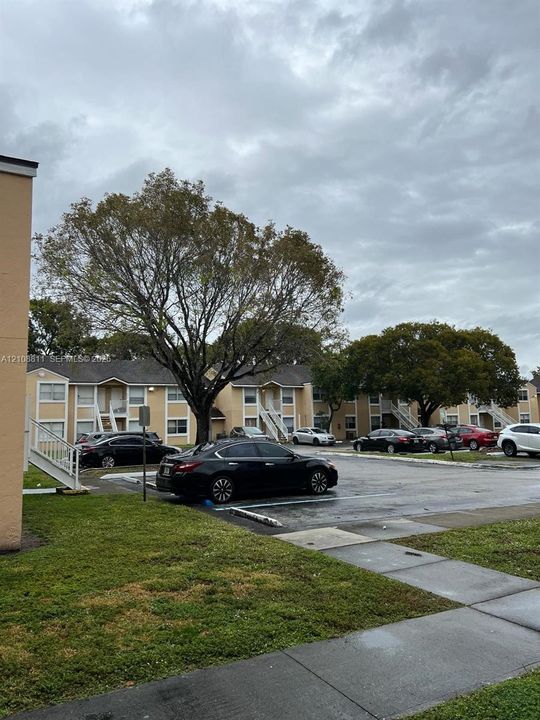 Two-story multi-unit structure featuring tan stucco exterior, shingled roofing, paved surface parking, and mature landscaping