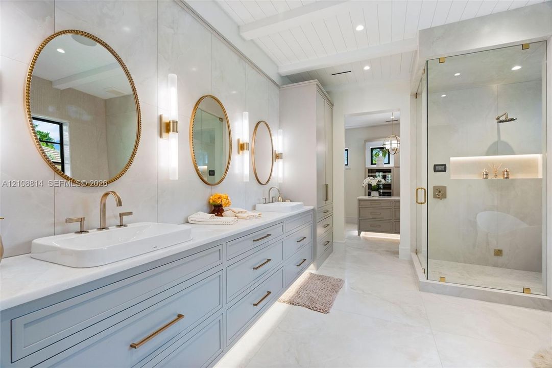 Dual-basin vanity featuring quartz countertops, large-format tile wall cladding, glass-enclosed walk-in shower with integrated recessed shelving, and custom cabinetry with gold-tone hardware