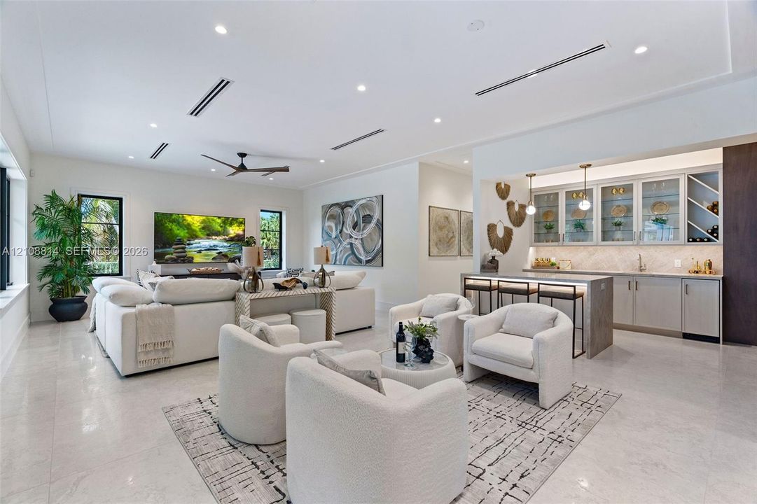 Open-concept living space featuring high ceilings with recessed lighting, large-format tile flooring, a dedicated wet bar with glass-front cabinetry, and a central island with integrated stone surfacing