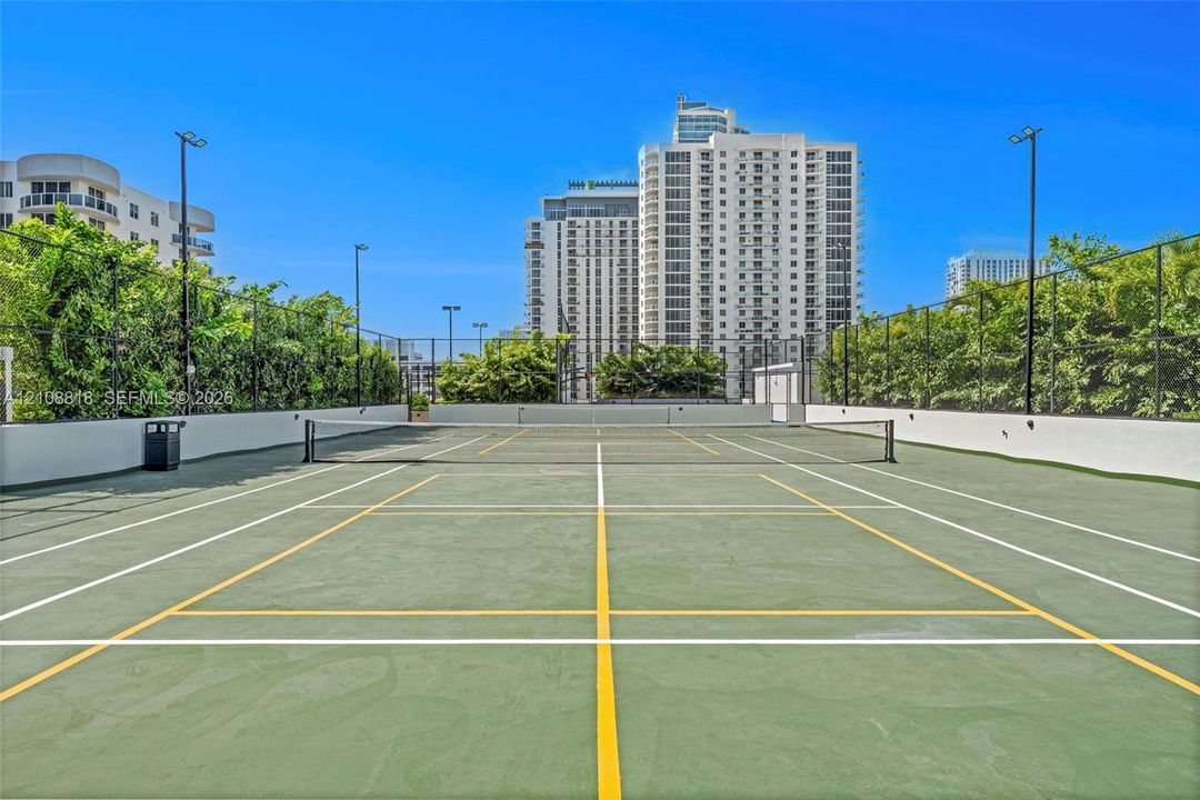 Hard-surfaced tennis court featuring perimeter chain-link fencing, high-mast lighting, and surrounding mature greenery