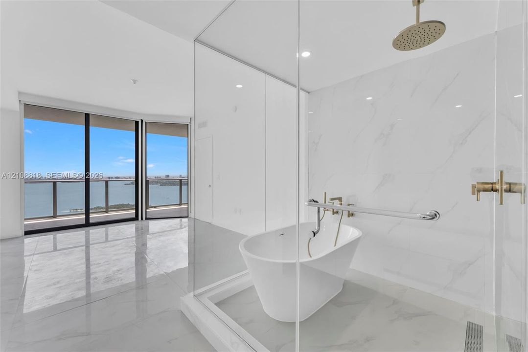 Freestanding soaking tub, glass-enclosed shower area with wall-mounted rainfall showerhead, polished stone-finish flooring, and floor-to-ceiling windows with water backdrop