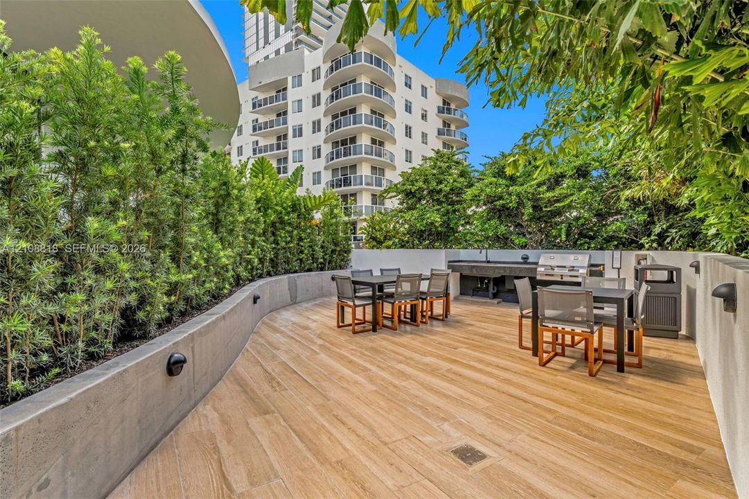 Outdoor terrace featuring wood-finish flooring, a built-in stone countertop with integrated grill, tiered concrete retaining wall, and landscape hedging