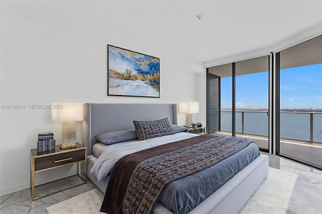 Floor-to-ceiling glass sliding doors, private balcony, neutral stone-finish flooring, and expansive water backdrop
