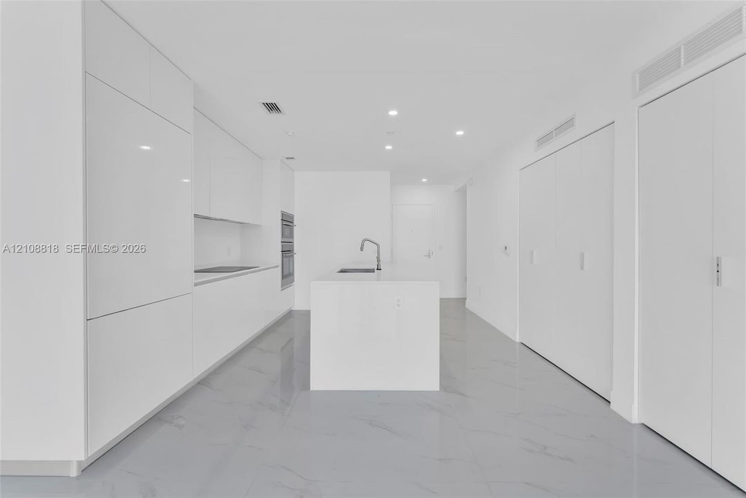 Minimalist kitchen featuring high-gloss cabinetry, a central island with a built-in sink, integrated double wall ovens, recessed ceiling lighting, and large-format polished flooring