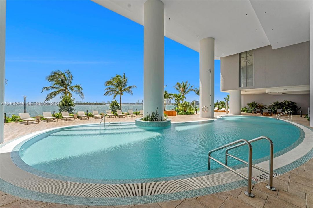 Curved swimming pool featuring tiled surrounds, stainless steel railings, structural columns, and paved deck flooring