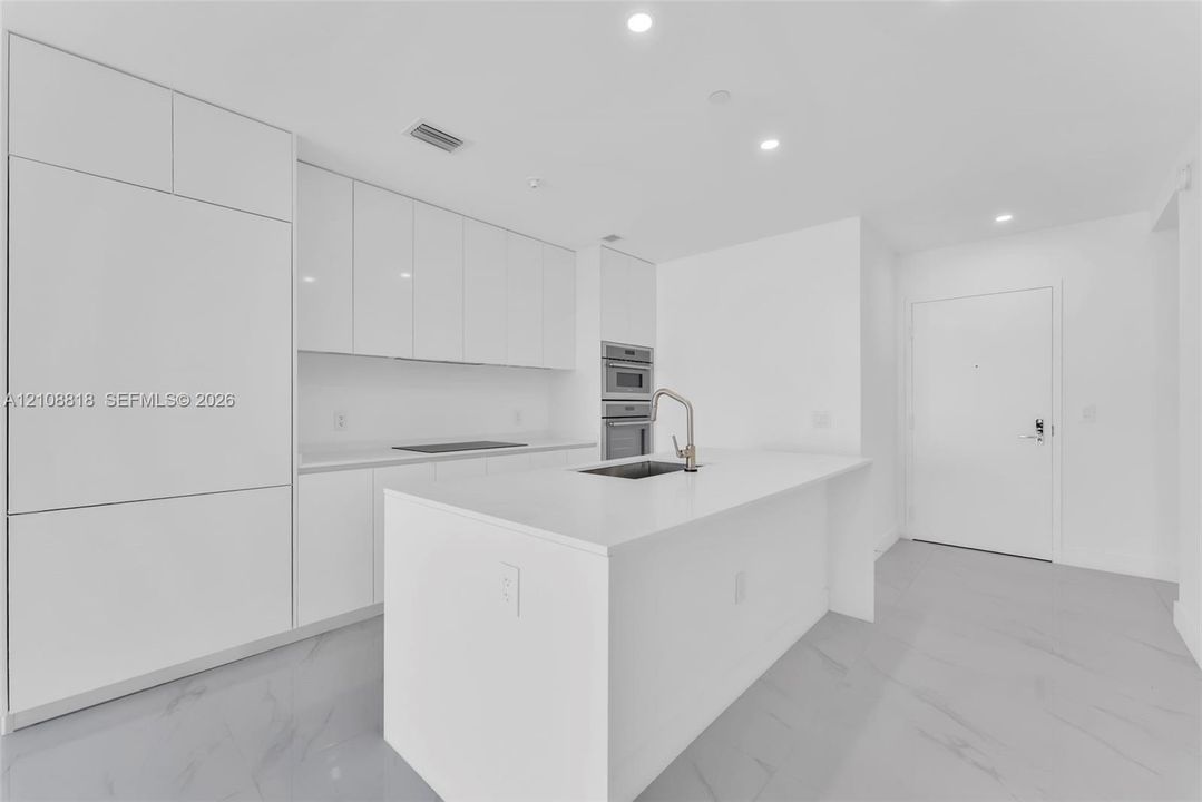Contemporary kitchen featuring white flat-panel cabinetry, a central island with a solid-surface countertop, an undermount sink with a gooseneck faucet, built-in stainless steel appliances, and large-format flooring