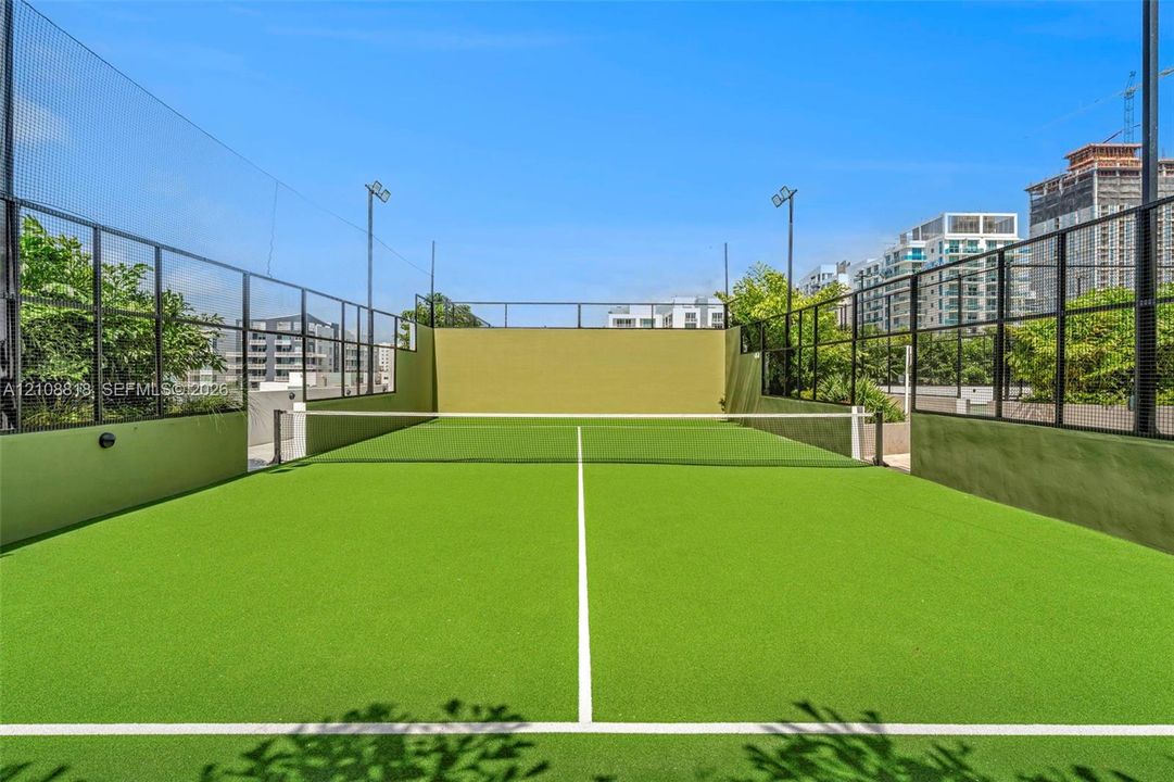 Paddle court featuring a synthetic turf surface, high-perimeter black metal fencing, integrated floodlighting, and solid concrete walls