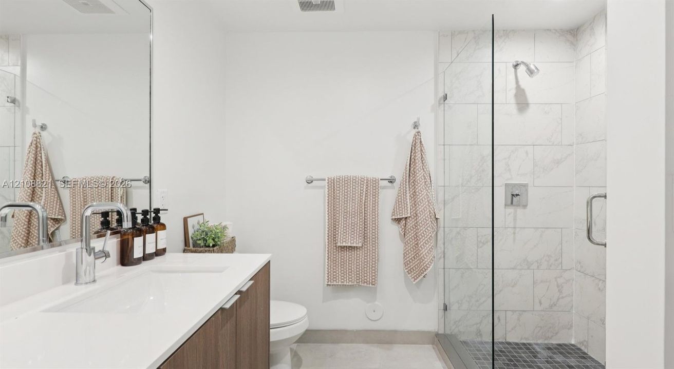 Floating wood-finish vanity with a white countertop and rectangular sink, oversized frameless mirror, glass-enclosed walk-in shower with tile surround, and polished chrome hardware