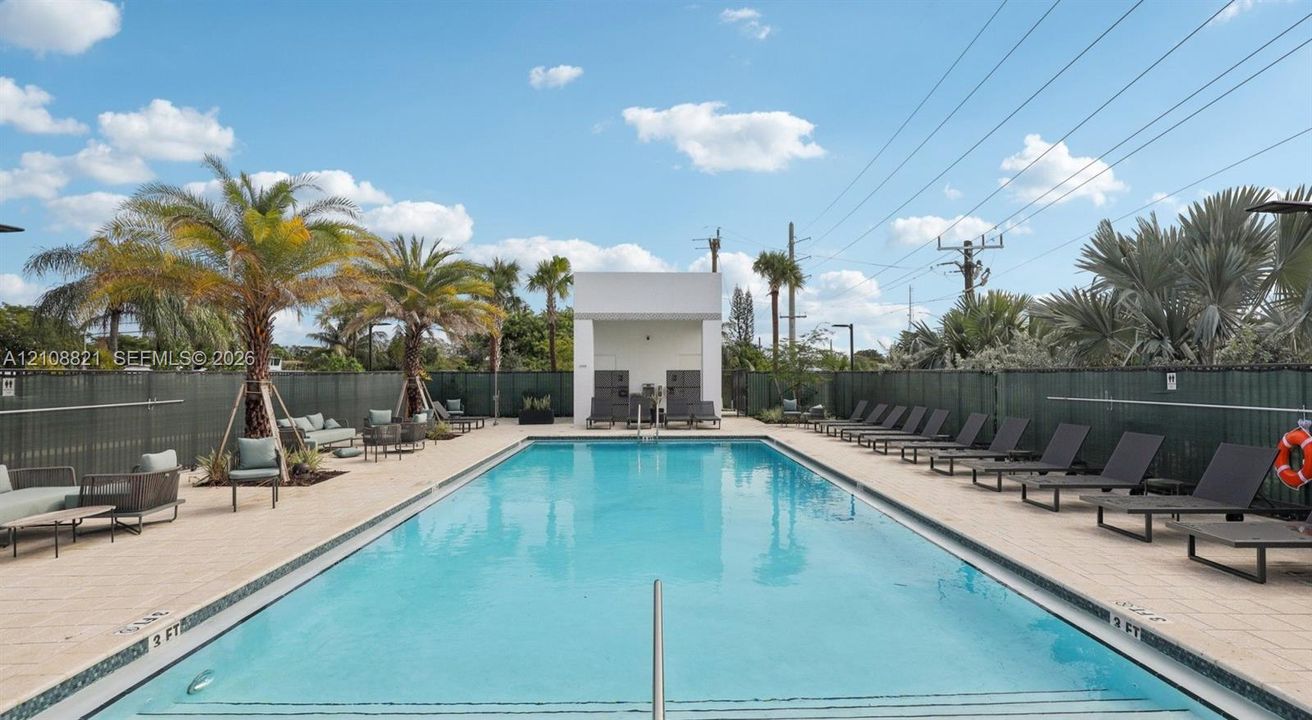 Rectangular swimming pool featuring a tiled deck, perimeter fencing, palm tree landscaping, and a central cabana structure