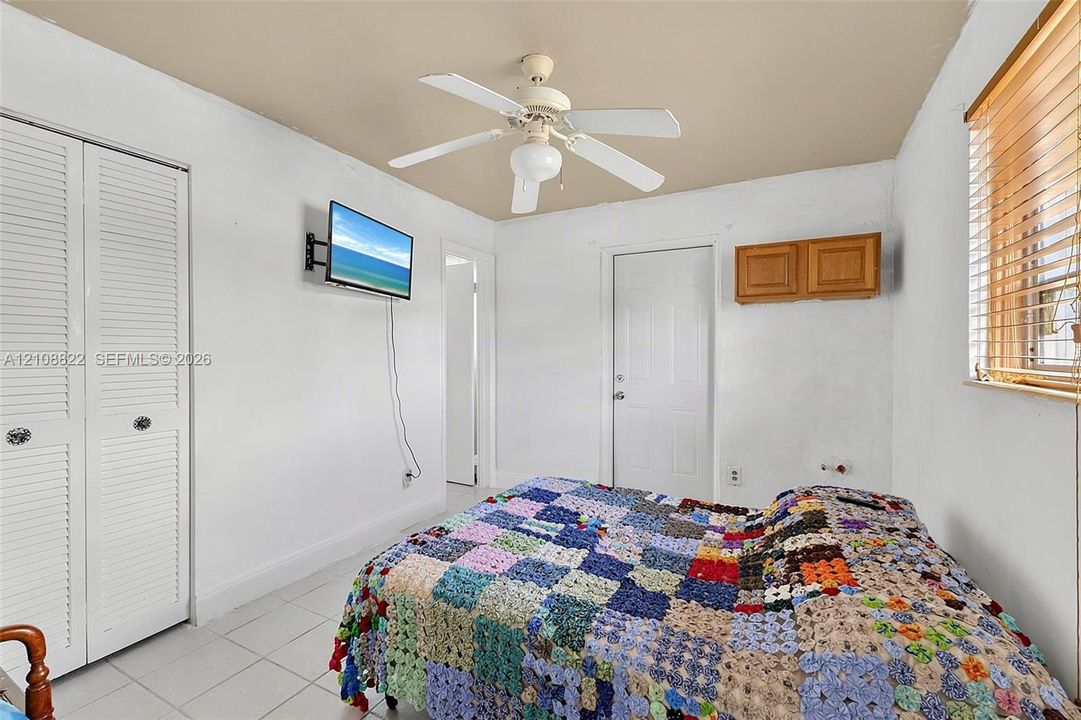 Bedroom featuring white louvered-panel closet doors, neutral-toned tile flooring, ceiling fan with integrated lighting, and wall-mounted wooden cabinetry