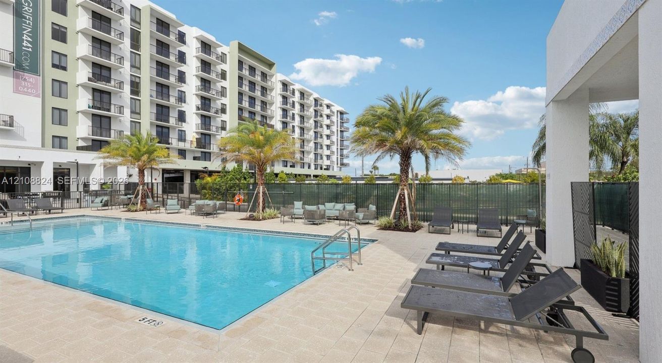 Outdoor swimming pool featuring a paved deck, surrounding palm trees, metal perimeter fencing, and concrete construction