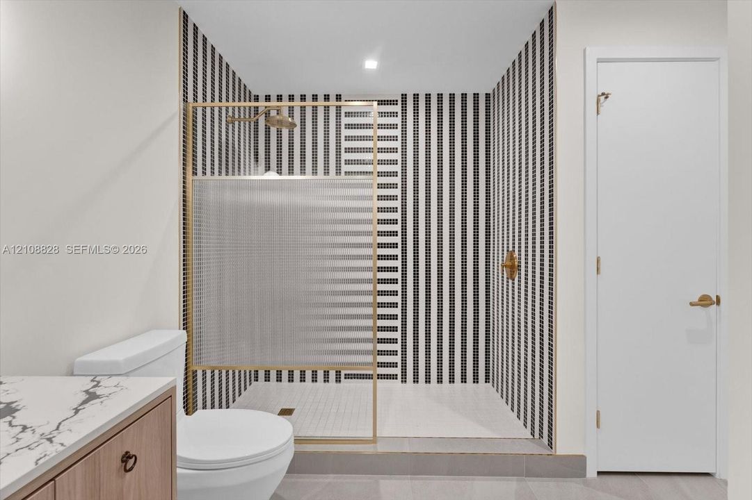 Walk-in shower featuring geometric black-and-white tile, brushed gold-finish hardware, glass panel enclosure, and stone-patterned vanity countertop