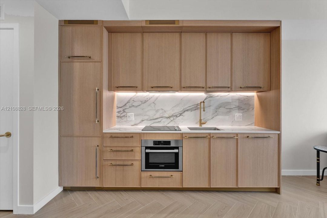 Custom flat-panel wood cabinetry with integrated refrigerator, stone slab backsplash, recessed under-cabinet lighting, brass-finish hardware, and herringbone wood-finish flooring