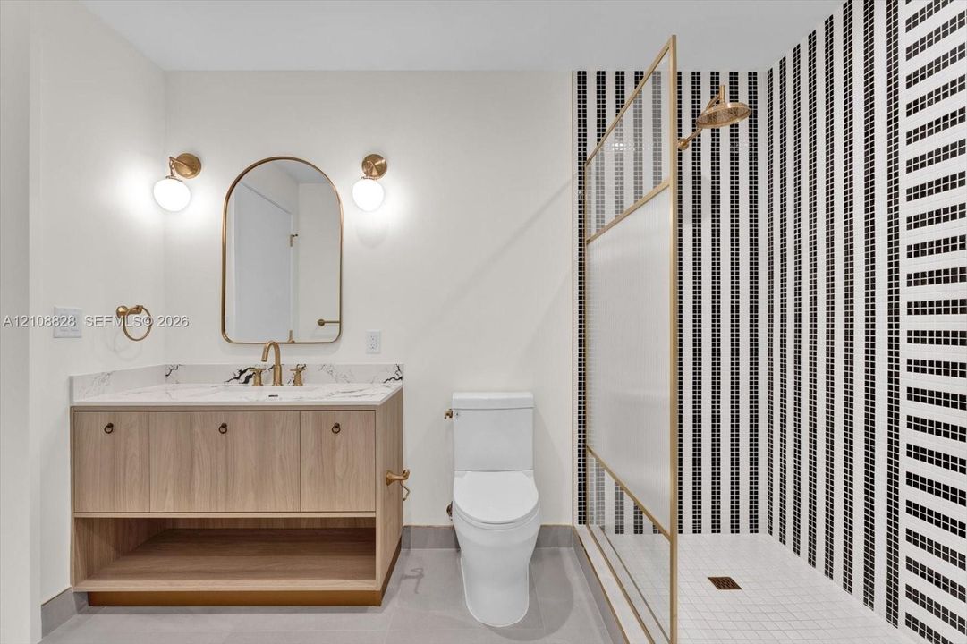 Bathroom featuring a wood-finish vanity with a stone-top counter, arched mirror, brass-toned fixtures, and a shower with a floor-to-ceiling geometric tile surround and textured glass enclosure