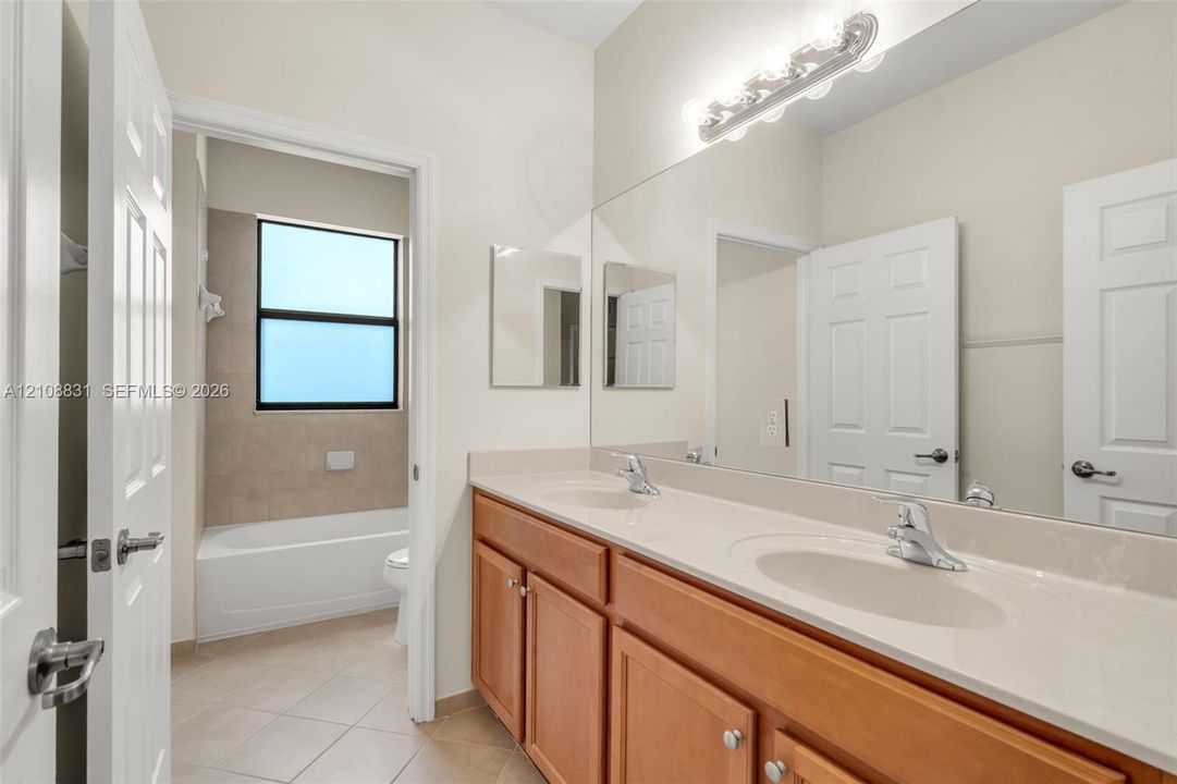 Dual-vanity cabinetry with an integrated countertop, full-width vanity mirror, polished chrome fixtures, tile flooring, and a tiled shower-tub surround