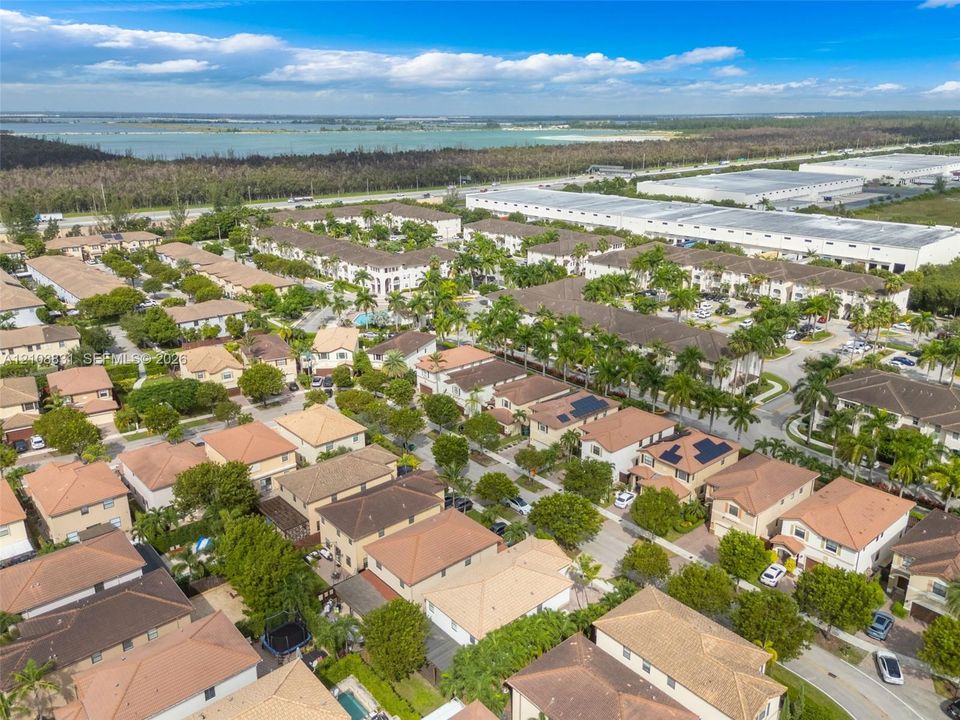 Aerial perspective of a residential community featuring multi-story dwellings with tile roofing, integrated solar panels, paved thoroughfares, and mature palm trees situated near a large body of water