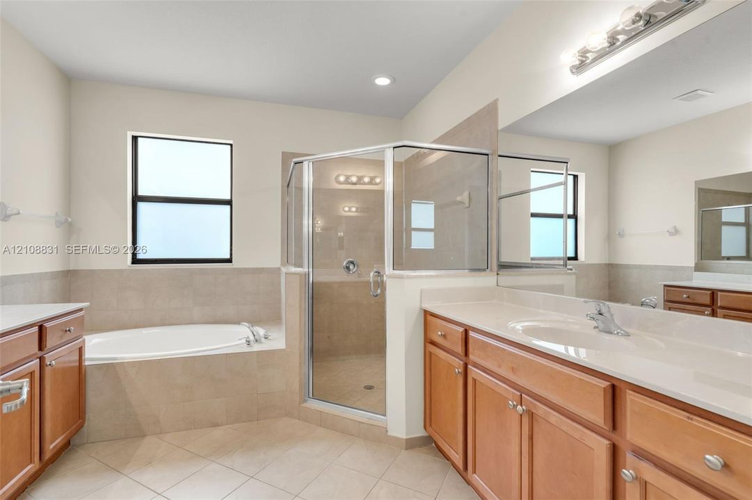 Bathroom featuring a soaking tub with tile surround, glass-enclosed shower, dual-sink vanity with light wood cabinetry, and neutral tile flooring