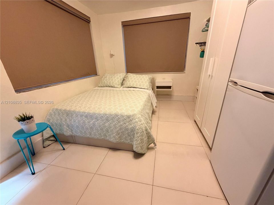 Large-format light-toned tile flooring, dual windows with roller shades, and a wall-mounted floating bedside shelf