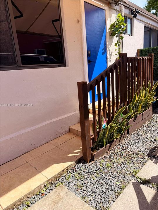 Exterior entrance featuring a solid-color door, tiled entryway, dark-toned wood railing, and stone ground cover