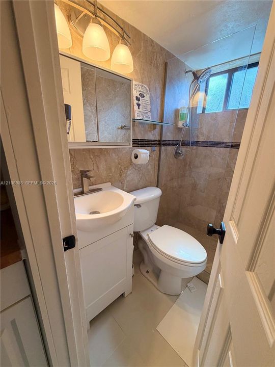 Bathroom featuring a pedestal sink with a single-handle faucet, stone-tile wall surround, glass shower panel, and three-light vanity fixture