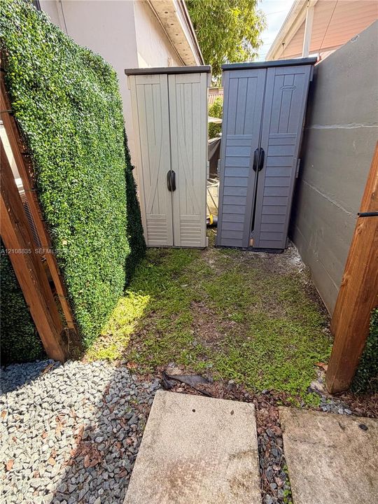 Exterior side yard featuring a concrete paver path, crushed stone surfacing, artificial turf privacy screening, and two vertical storage units