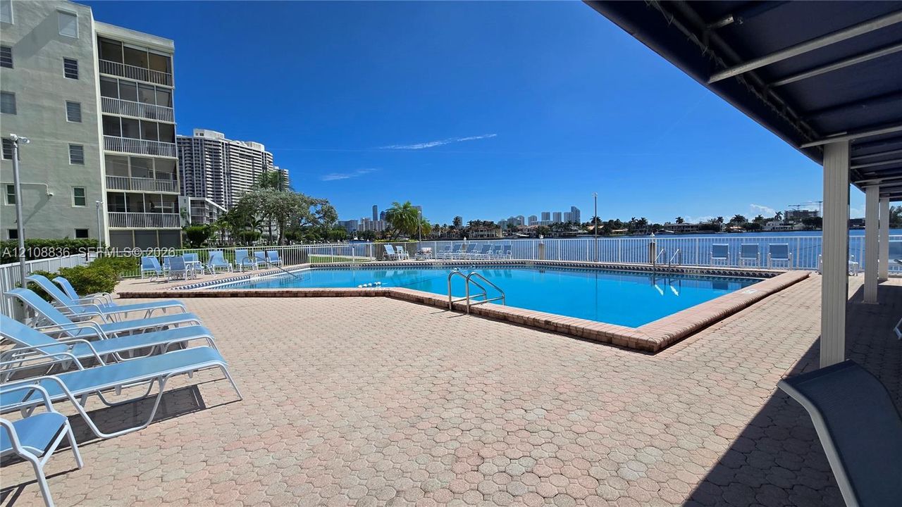 Waterfront swimming pool featuring paver decking, white metal safety fencing, and an overhead canopy structure