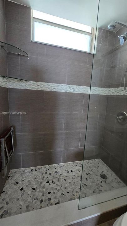 Walk-in shower featuring large-format wall tile, a horizontal decorative mosaic tile accent, pebble-style floor tiling, a glass enclosure, and a transom window
