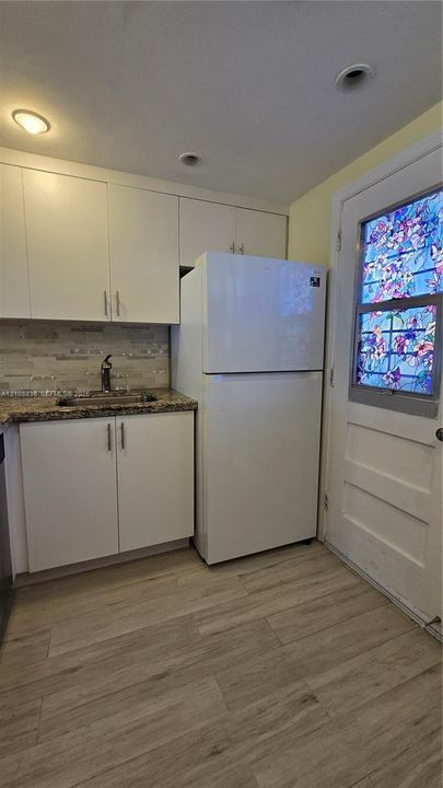 Kitchenette featuring white shaker-style cabinetry, stone-surface countertops, textured tile backsplash, wood-finish flooring, and a decorative stained-glass panel door