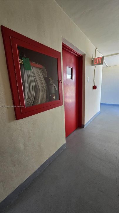 Hallway featuring a painted metal door, wall-mounted fire safety cabinet with glass panel, and illuminated exit signage