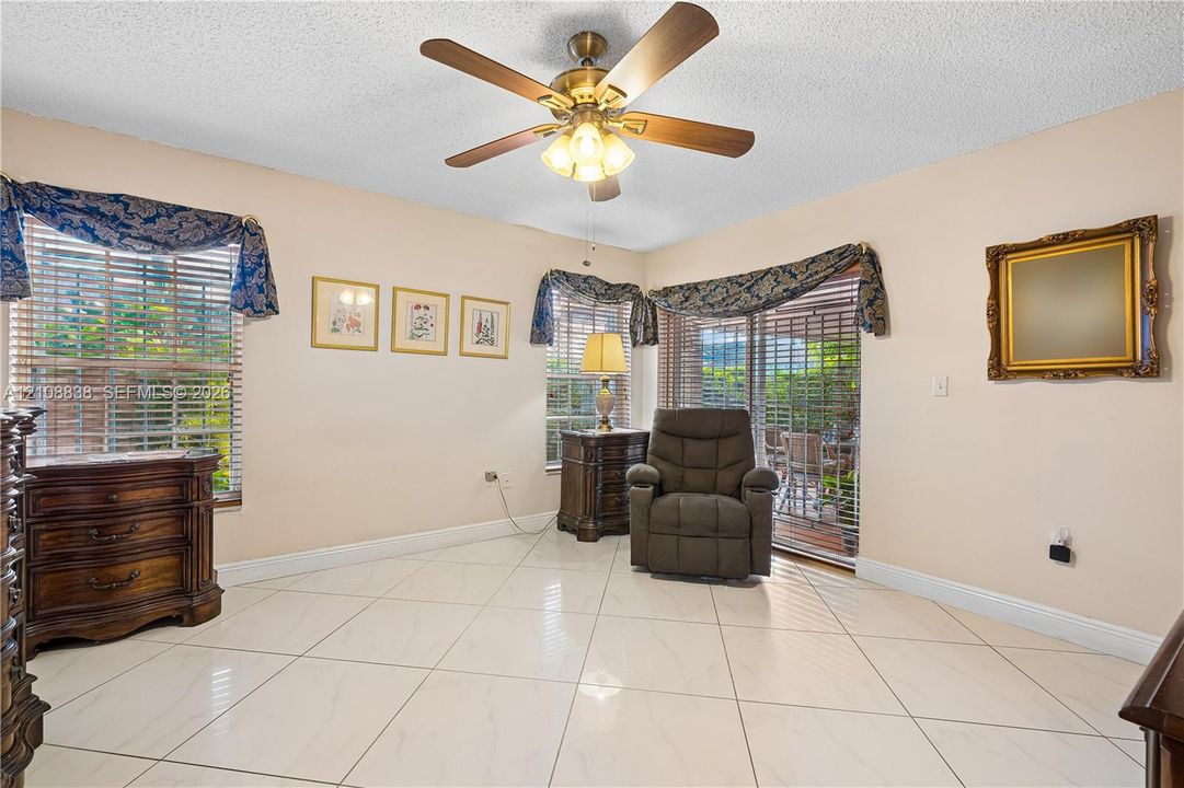Large-format light-toned tile flooring, multi-pane windows with horizontal blinds, central ceiling fan with integrated lighting, and glass sliding door providing outdoor access
