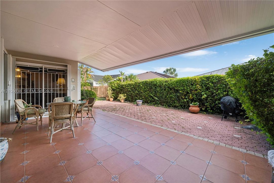 Covered patio featuring ceramic tile flooring, retractable awning, and a brick-paver hardscaped area enclosed by a mature hedge