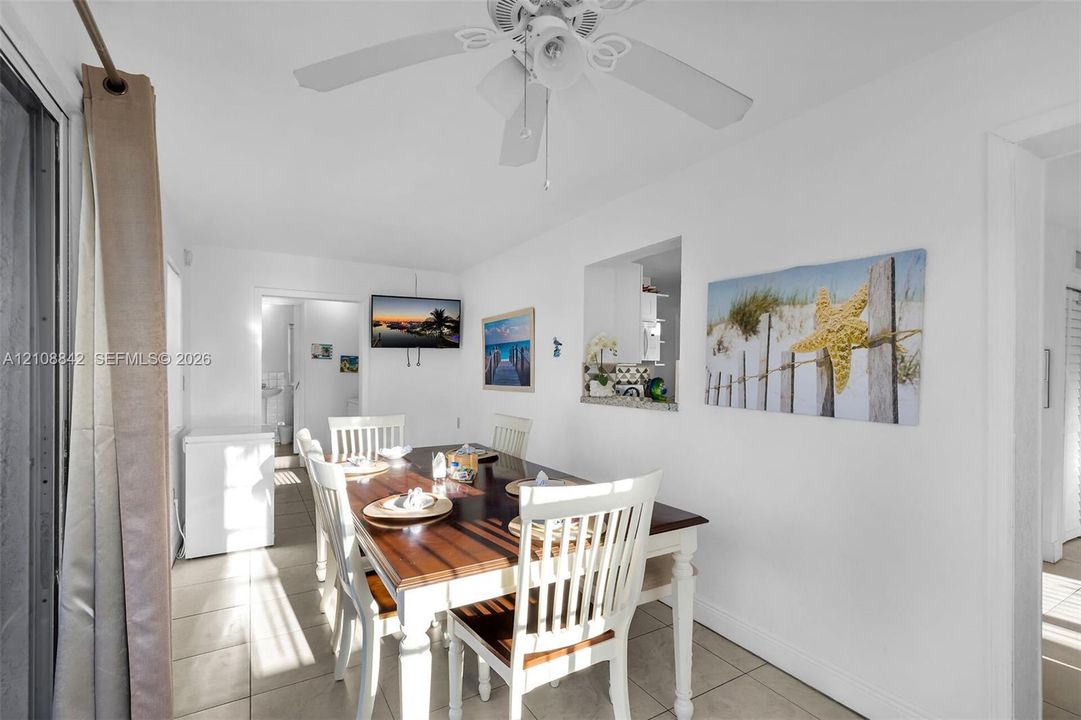 Dining area featuring light-toned tile flooring, a ceiling fan with integrated lighting, and a wall cutout providing a sightline to the kitchen
