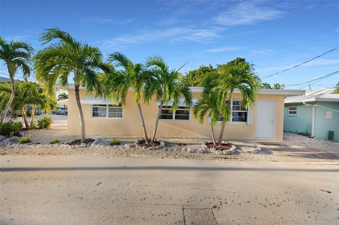 Single-story exterior featuring light-toned stucco siding, white window frames with metal awnings, and mature palm trees within a stone-mulched landscape