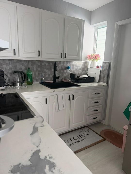 White cabinetry with matte black hardware, stone-finish countertops, hexagonal tile backsplash, matte black kitchen faucet, and integrated cooktop