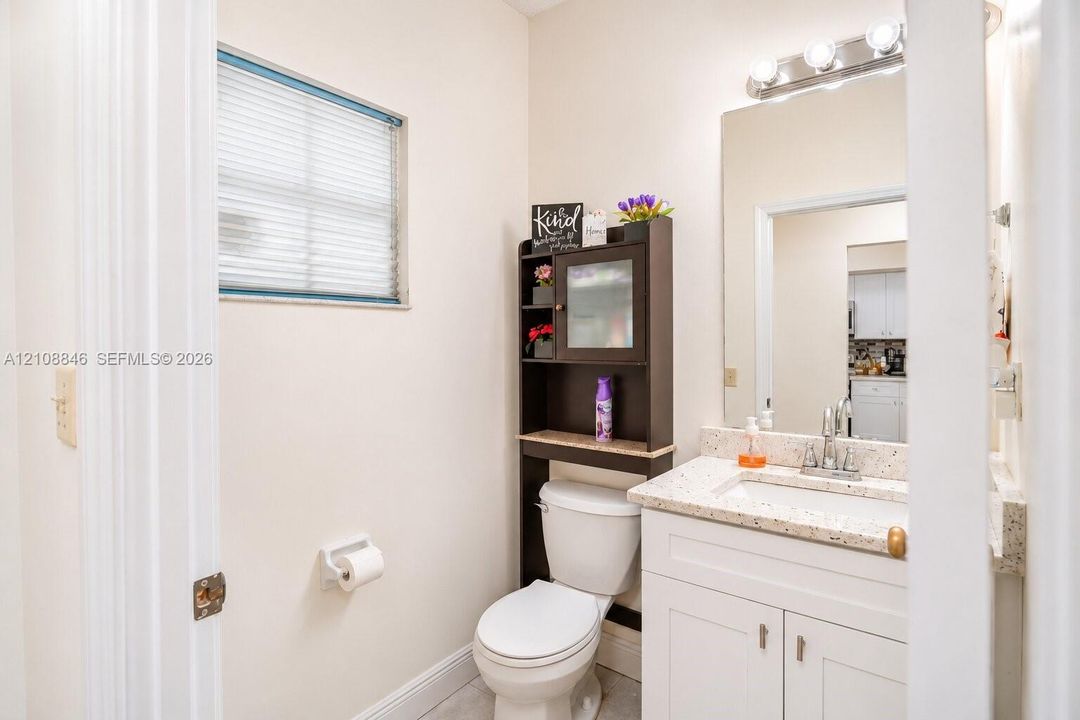 Bathroom featuring a vanity with a white cabinet and stone-composite countertop, integrated sink, wall-mounted mirror, four-bulb light fixture, and horizontal blind window