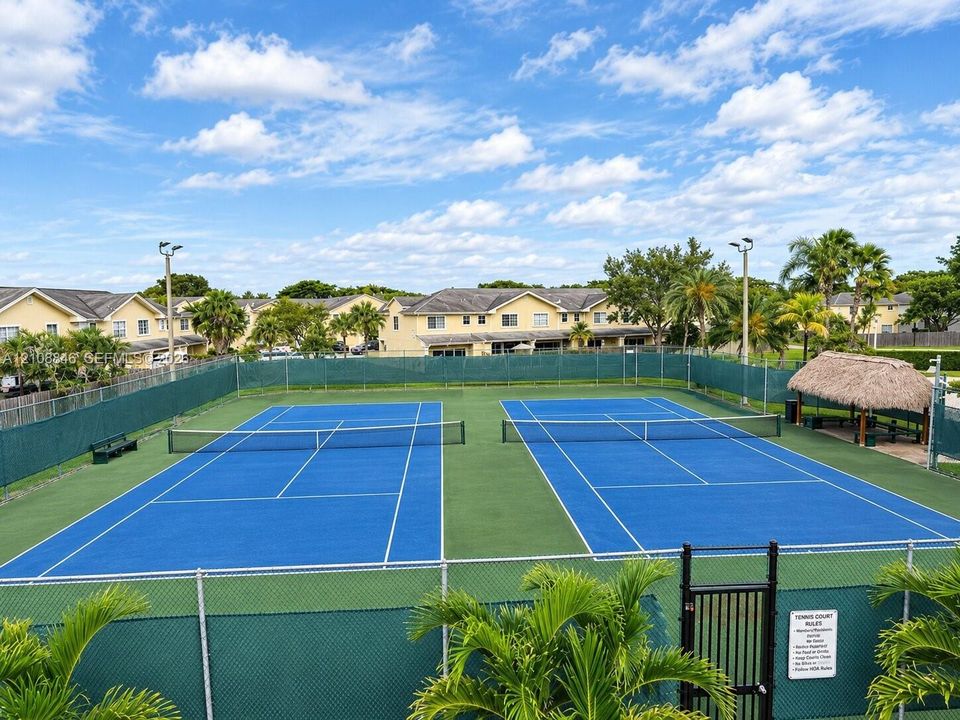 Dual tennis courts featuring specialized surface markings, perimeter chain-link fencing, stadium-style lighting, and a thatched-roof pavilion
