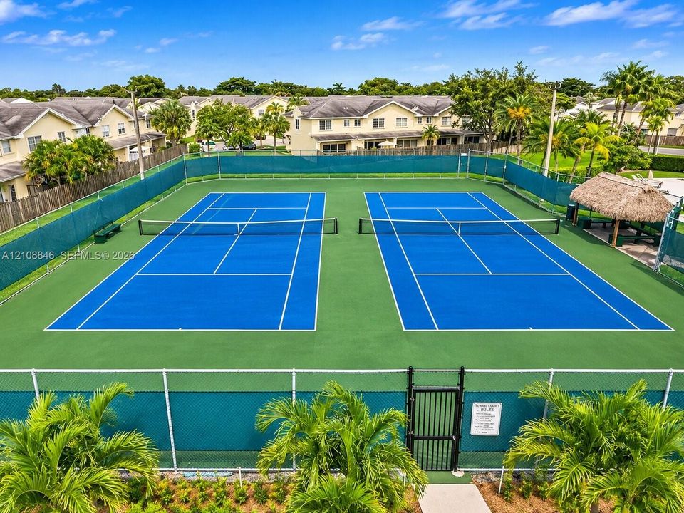 Dual tennis courts featuring professional surfacing, perimeter chain-link fencing, and a thatch-roofed cabana