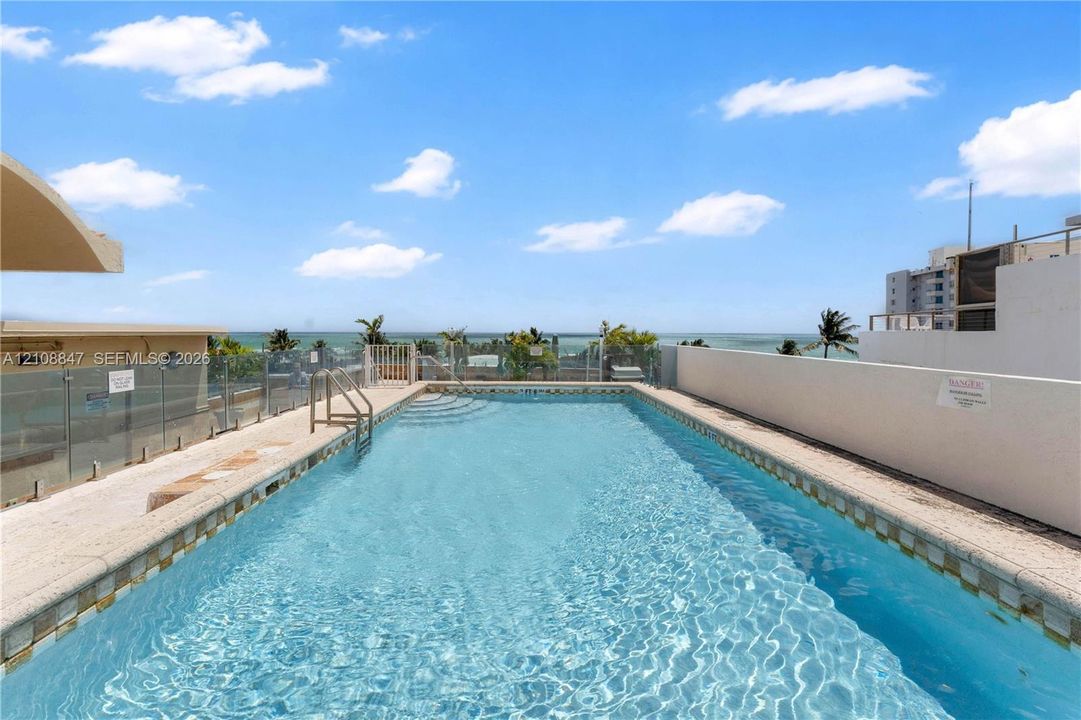 Rooftop swimming pool featuring stone-style decking, a metal ladder, and glass panel railing