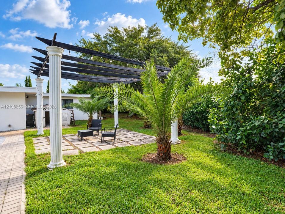 Outdoor living area featuring a pergola supported by classic columns, stone paver patio, manicured lawn, and mature landscaping