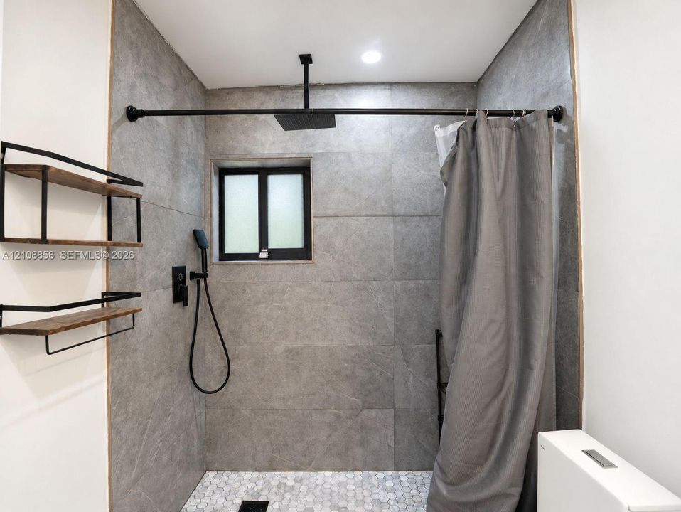 Shower area featuring large-format grey tile walls, a white hexagonal tile floor, matte black rainfall showerhead with handheld sprayer, and built-in wood-finish shelving with black metal framing