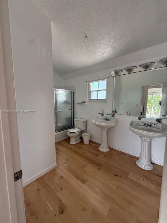 Bathroom featuring wood-finish flooring, dual pedestal sinks, a large wall-mounted mirror with integrated lighting, and a glass-enclosed shower