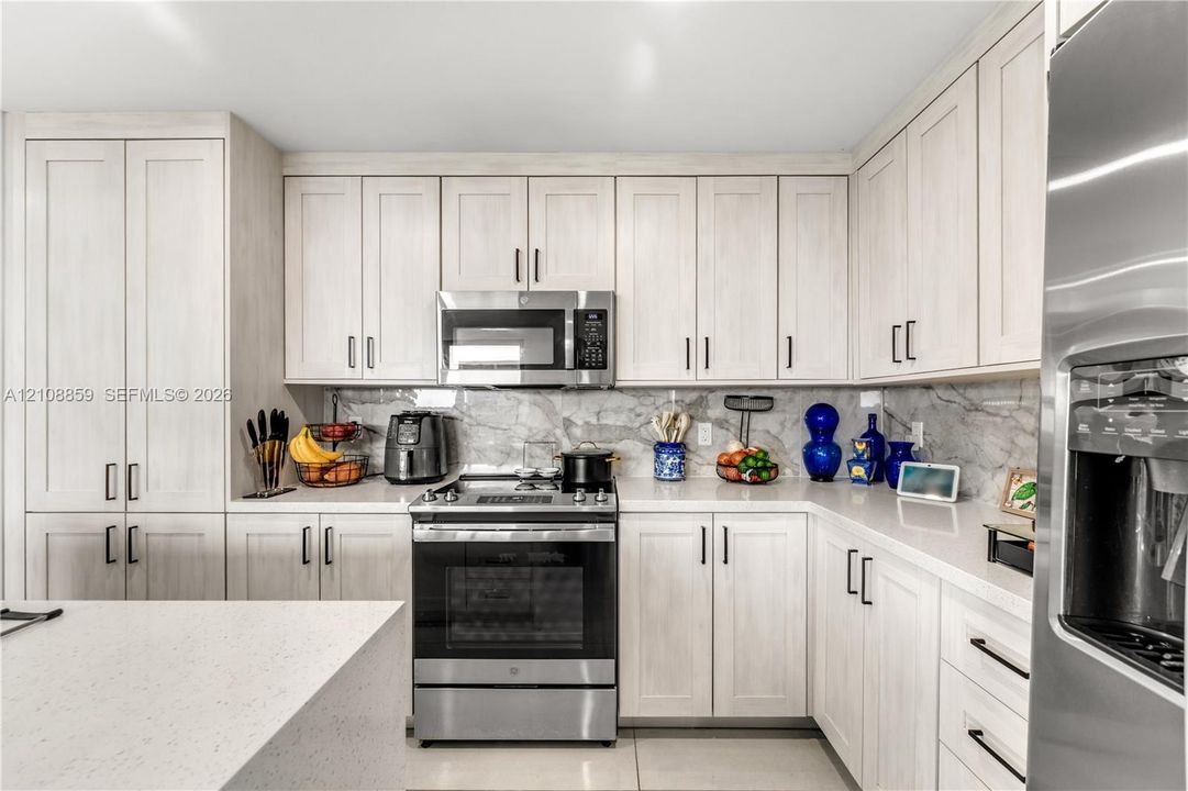 Kitchen featuring light-toned shaker cabinetry, stone-finish backsplash, quartz countertops, stainless steel appliances, and matte black hardware