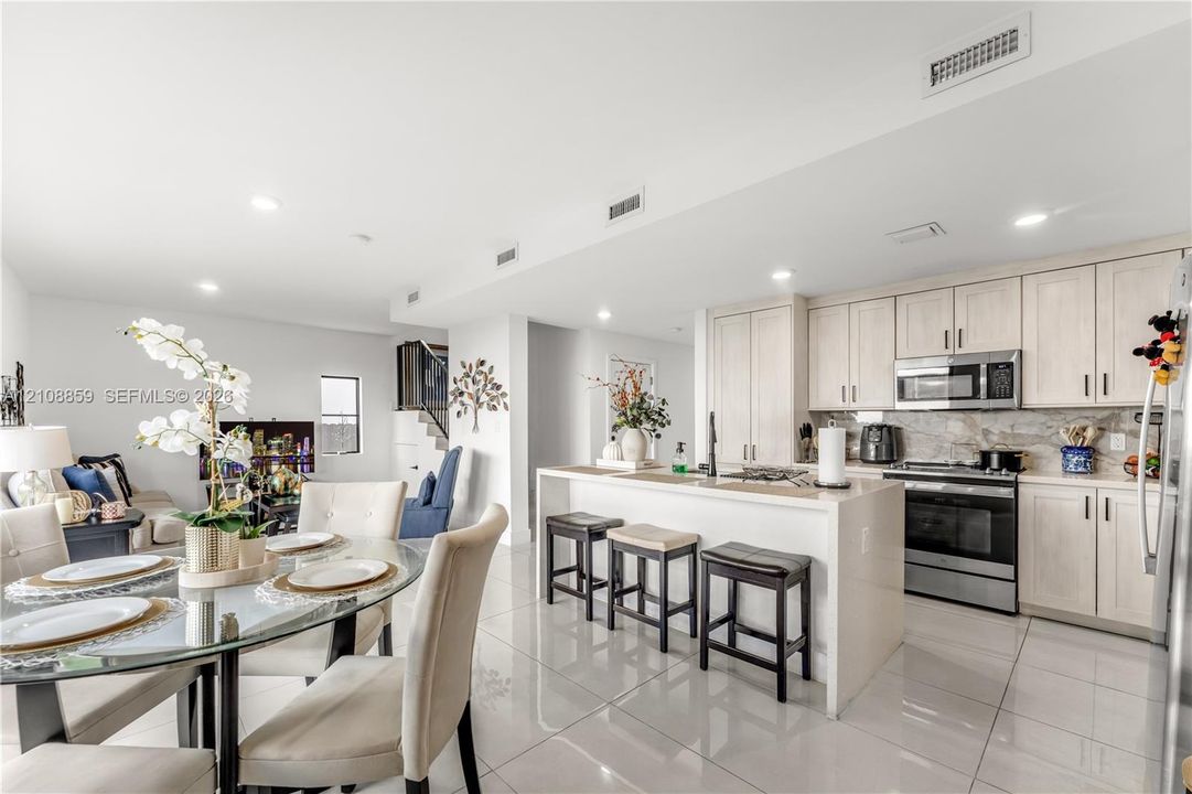 Open-concept kitchen featuring light-toned cabinetry, stainless steel appliances, a stone-finish backsplash, a central island with integrated seating, and high-gloss tile flooring