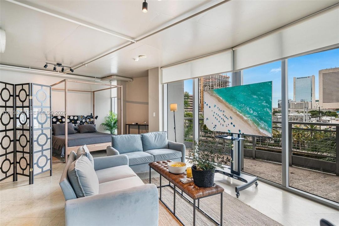 Floor-to-ceiling windows, light-toned stone-finish flooring, industrial-style track lighting, and a private balcony with metal railing