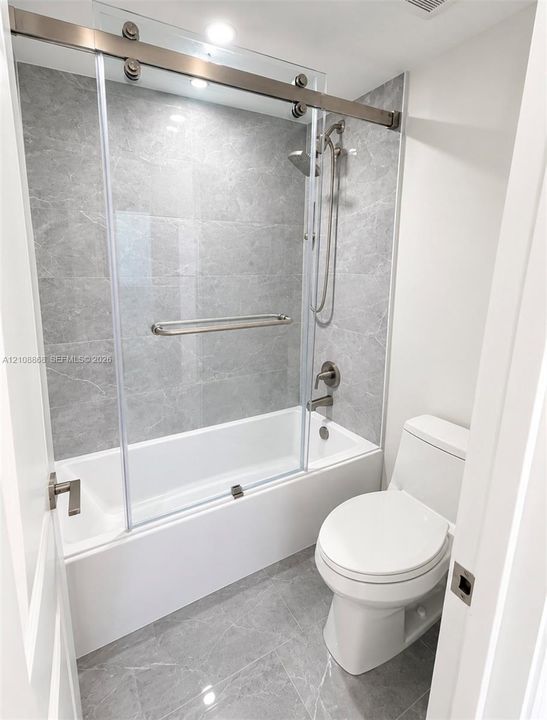 Full bathroom featuring large-format grey tile flooring, a tub-shower combination with a sliding glass door, grey tile surround, and a chrome hand-held shower fixture