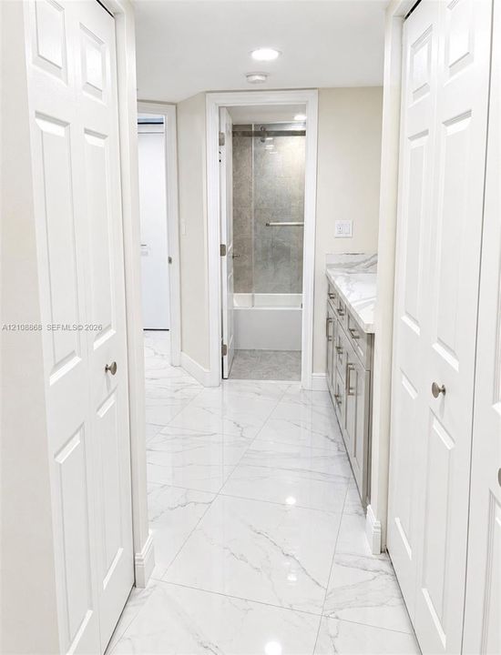 Bathroom featuring large-format stone-finish flooring, a vanity with stone-finish countertop, dual bi-fold closet doors, and a shower-tub combination with a glass enclosure