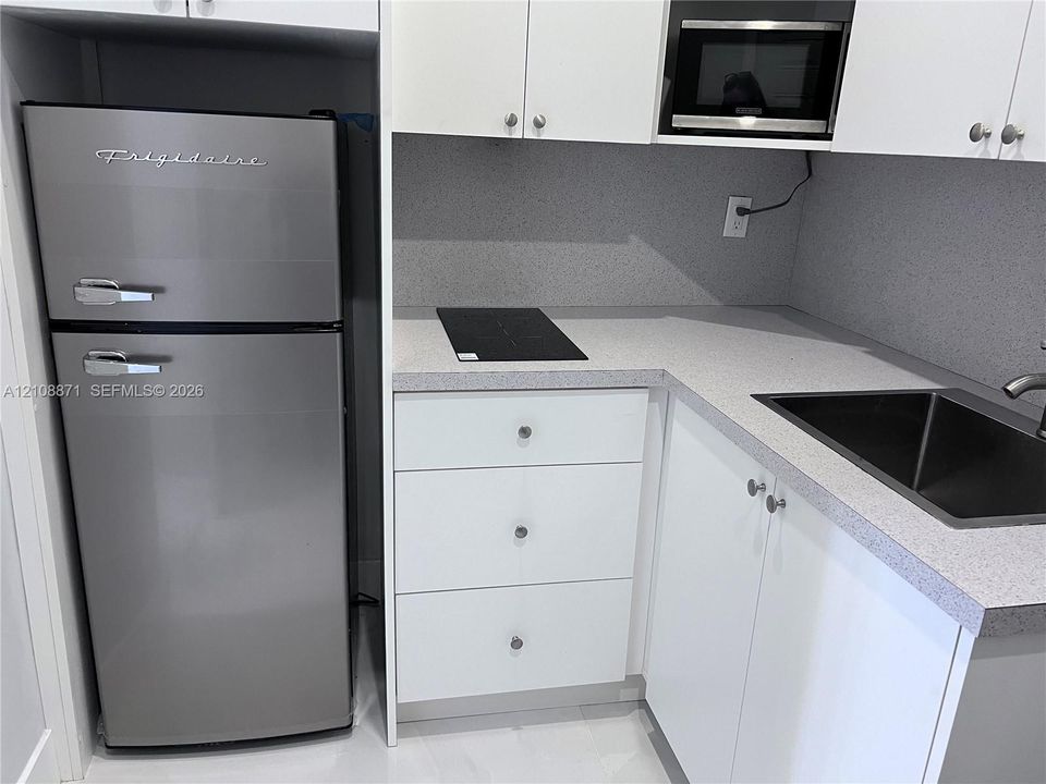 Compact kitchen featuring white cabinetry, stone-finish countertops, a stainless steel refrigerator, an integrated cooktop, and a built-in microwave