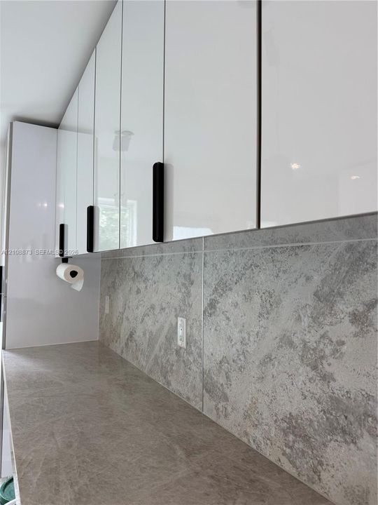 High-gloss white cabinetry featuring matte black hardware, textured stone-finish backsplash, and solid-surface countertop