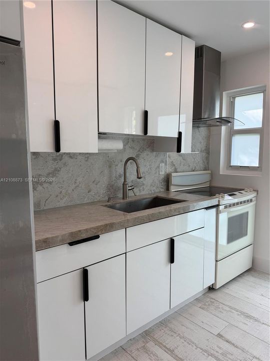 Glossy white cabinetry featuring matte black hardware, stone-finish countertops, coordinating backsplash, stainless steel sink with high-arc faucet, and wood-finish tile flooring